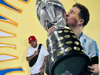 AB Quintanilla is posing next to a mural of Lionel Messi lifting Copa America trophy.